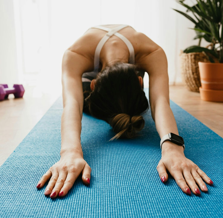 Person macht Yoga-Stretch auf blauer Matte, Arme nach vorn gestreckt, Pflanzen und Hanteln im Hintergrund.